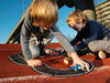 Two young children play with the Large Flexible Toy Race Track - Grand Prix on an outdoor running track under bright sunlight, with buildings and other structures visible in the background.