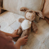 A child reaches for a soft Olli Ella Cozy Dinkum - Dog Digby doll, dressed in a dog outfit, lying on a textured beige blanket with a cushion in the background.
