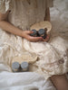 A child in a lacy beige dress holds a Wooden Mom and Baby Elephant Push Toy Set. Another similar toy rests in her lap. The scene is soft and serene, with the child sitting on a plush white surface. The elephants, designed to enhance fine motor skills, have simple designs with gray wheels and are free of harmful chemicals.