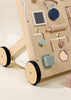 The Wooden Activity Walker features black wheels and a variety of interactive shapes, knobs, and cutouts designed as sensory toys for young children. The background is a light, neutral color.