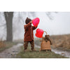 A young child in a brown coat and hat holds two red heart-shaped balloons on a misty path, beside a wicker basket with the GOTS-certified Senger Naturwelt Stuffed Animal - Large Brown Bear and an attached balloon.