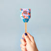 A hand with purple nail polish holds a Garden Seed Pop (Single) with a colorful wrapper labeled "Strawberry Seed Pop" and "Plant Me!"—a playful touch for any garden kit—set against a plain light blue background.