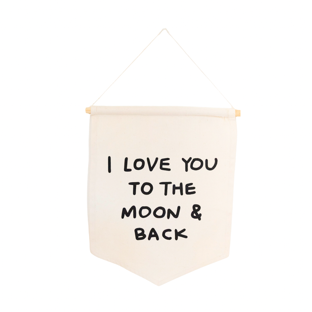 The I Love You To The Moon And Back Canvas Sign features a cream fabric hanging from a wooden rod with string, displaying “I LOVE YOU TO THE MOON & BACK” in bold, black hand-lettered text.