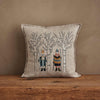 The Coral & Tusk Aspen Skiers Pillow is a square beige cushion with embroidered animals in winter attire among bare trees, perfect for cozy winter decor on a neutral wall and wooden surface.