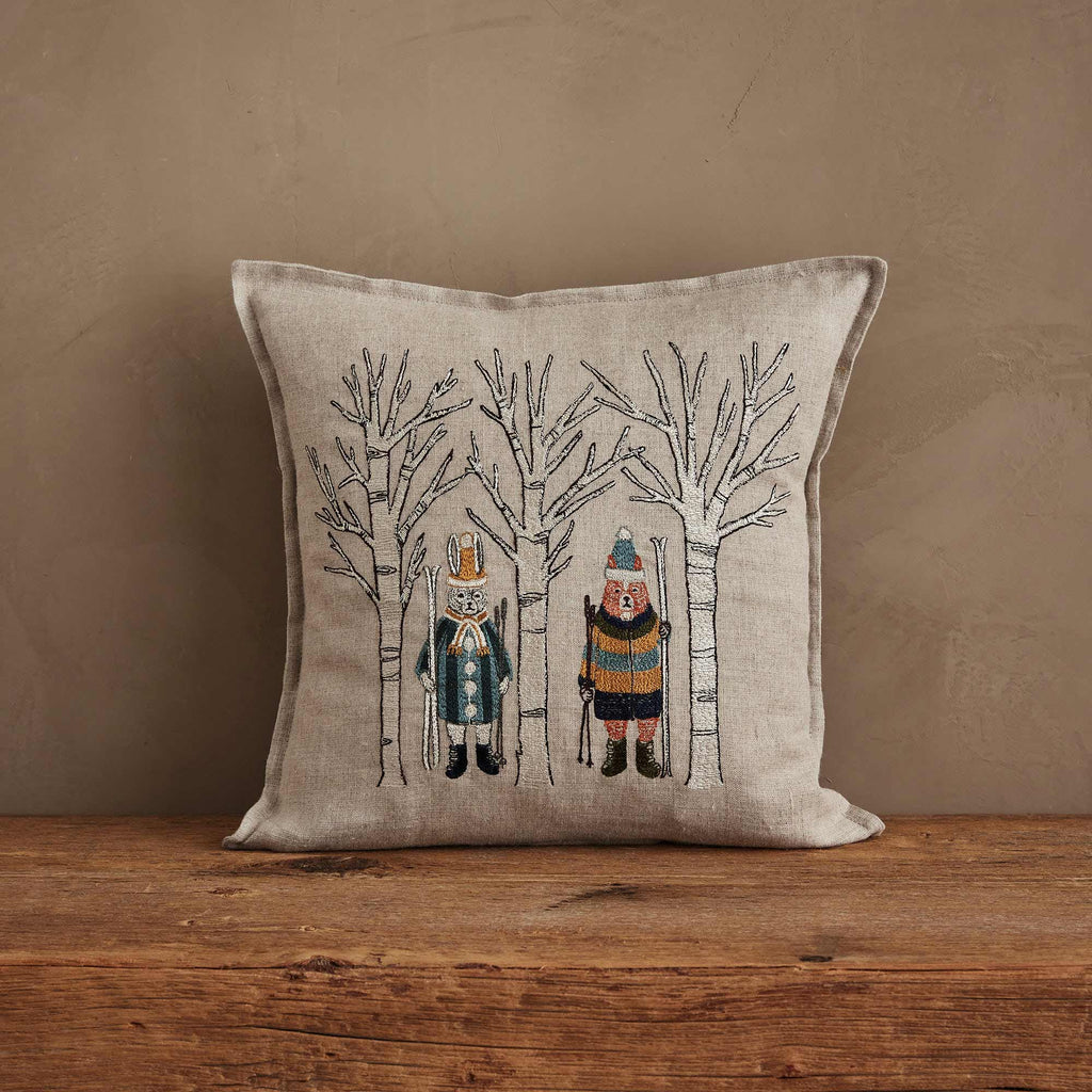 The Coral & Tusk Aspen Skiers Pillow is a square beige cushion with embroidered animals in winter attire among bare trees, perfect for cozy winter decor on a neutral wall and wooden surface.