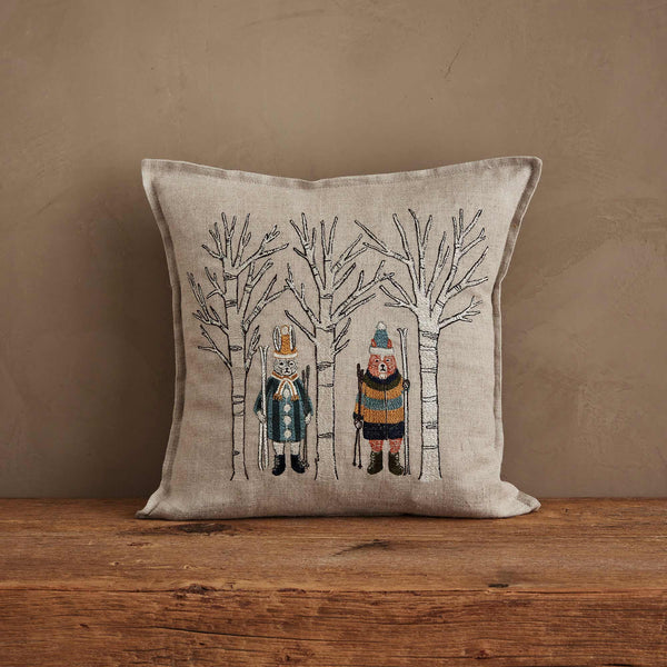 The Coral & Tusk Aspen Skiers Pillow is a square beige cushion with embroidered animals in winter attire among bare trees, perfect for cozy winter decor on a neutral wall and wooden surface.