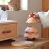 A hand lifts the lid of the Christmas Tree Storage Box, shaped like a smiling tree, next to another wooden box with a face and a bottle of perfume on a light wood table.
