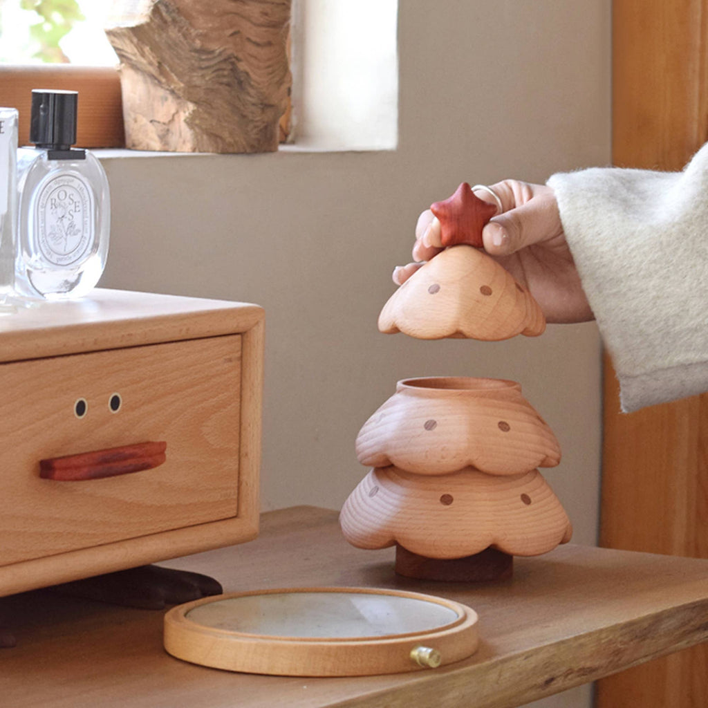 A hand lifts the lid of the Christmas Tree Storage Box, shaped like a smiling tree, next to another wooden box with a face and a bottle of perfume on a light wood table.