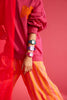 A person in a pink sweater and orange-striped pants stylishly showcases four Mini Kyomo Watch - Lavender Field pieces on their arm against a pink background. Their face is hidden; the pose is dynamic and eye-catching.