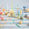 Assorted cupcakes with colorful icing and playful toppers like dinosaurs and mushrooms on a striped background, adorned with Meri Meri Dinosaur Candles and festive decorations.