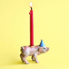 A whimsical Pig Cake Topper with a red candle burning on its back against a plain yellow background. The pig is decorated with a blue and white striped party hat.