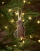 The Coral & Tusk Bear with Tinsel Ornament, featuring a fabric bear with tree designs, hangs from a Christmas tree branch amid glowing lights—a charming woodland accent for your holiday home decor.