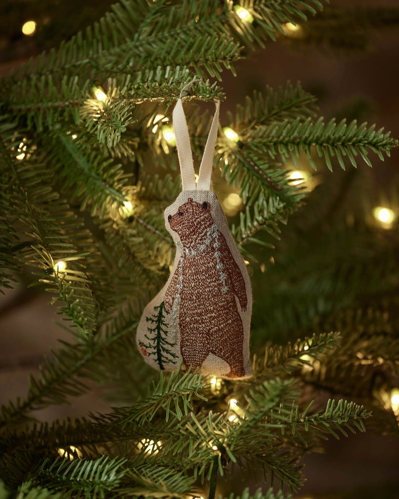 The Coral & Tusk Bear with Tinsel Ornament, featuring a fabric bear with tree designs, hangs from a Christmas tree branch amid glowing lights—a charming woodland accent for your holiday home decor.