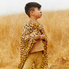 A young child with drawn-on whiskers stands in a dry grassy field, wearing Sarah's Silks Animal Print Playsilk - Cheetah as a cape over tan clothing, looking off to the side.