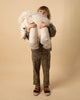 A young child stands against a beige backdrop, holding a large, BigStuffed Albino Mammoth Stuffed Animal. The child is dressed in a matching outfit with a leopard print pattern and is wearing maroon shoes. The child looks directly at the camera, partially obscured by the mammoth plush.