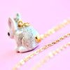 A close-up of a cute, small Goldentail Rabbit Necklace featuring a hand-painted bunny charm with a 24k gold plated tail attached to a delicate gold chain, set against a soft pink background. The charm displays intricate details, giving the bunny a realistic appearance.