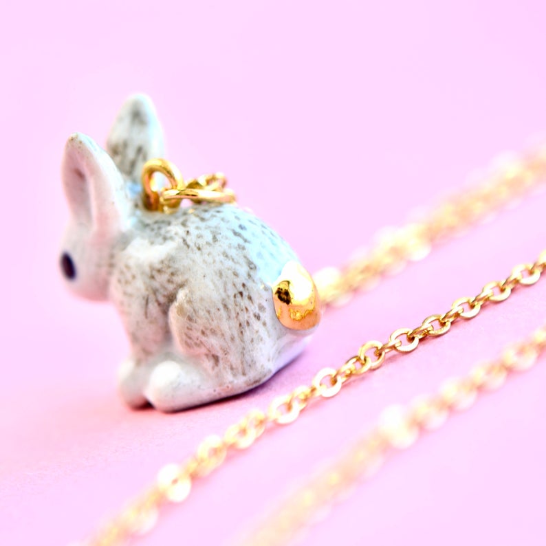 A close-up of a cute, small Goldentail Rabbit Necklace featuring a hand-painted bunny charm with a 24k gold plated tail attached to a delicate gold chain, set against a soft pink background. The charm displays intricate details, giving the bunny a realistic appearance.