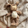 An Olli Ella Cozy Dinkum - Dog Digby plush toy with soft brown fur, floppy ears, closed eyes, and a furry white belly with a small rainbow, rests on a textured beige blanket. Made from recycled materials.