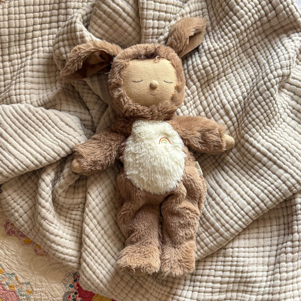 An Olli Ella Cozy Dinkum - Dog Digby plush toy with soft brown fur, floppy ears, closed eyes, and a furry white belly with a small rainbow, rests on a textured beige blanket. Made from recycled materials.