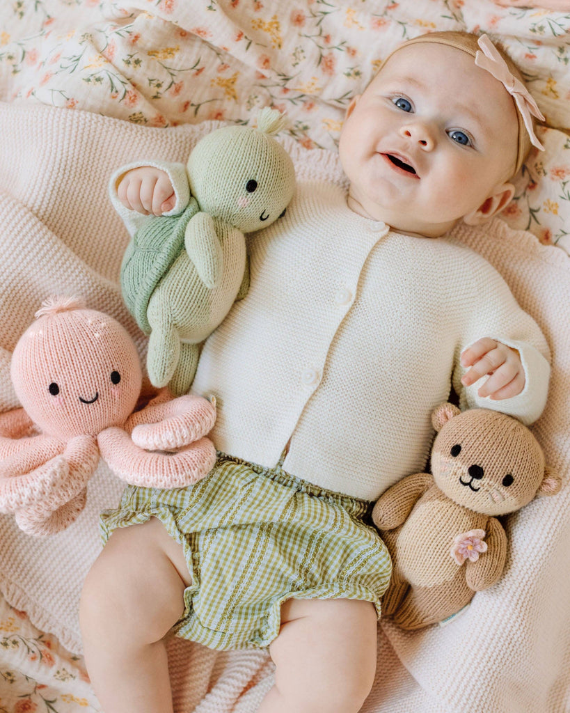 A smiling baby lies on a soft floral blanket, wearing a cream cardigan, green checkered shorts, and a pink bow headband, holding the heirloom-quality hand-knit Cuddle + Kind Baby Sea Turtle in Kelp Green.