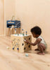 A young child in a beige romper crouches on a wooden floor, playing with the Wooden Activity Cube, a toddler toy featuring beads, shapes, and maze games that help develop fine motor skills in a softly lit minimalist room.