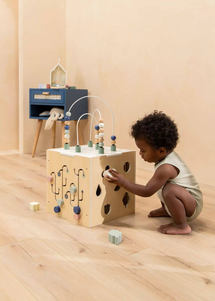 A young child in a beige romper crouches on a wooden floor, playing with the Wooden Activity Cube, a toddler toy featuring beads, shapes, and maze games that help develop fine motor skills in a softly lit minimalist room.