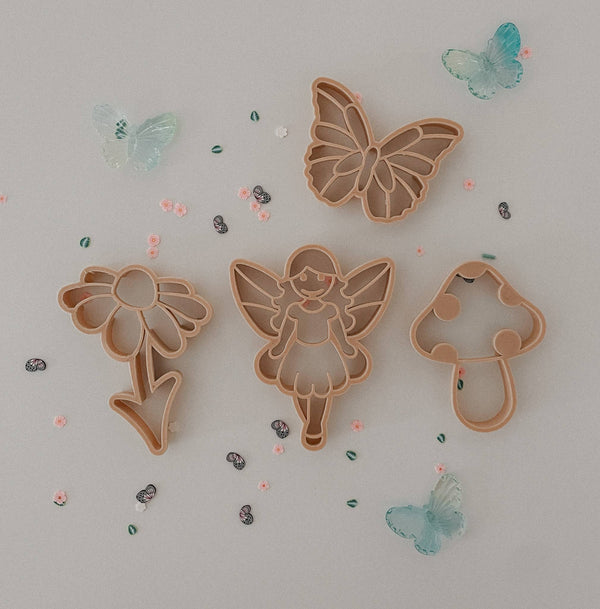 The Fairy Cutter Set - Eco Playdough Tool features four imaginative shapes—a flower, fairy, butterfly, and mushroom—beautifully arranged on a light surface and accented with decorative plastic butterflies and small beads, creating the perfect fairytale-inspired cutter set for sensory play.