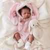 A baby in a pink hand-knit outfit with bunny ears lies on a soft cream blanket, cuddling the Cuddle + Kind Baby Bunny - Confetti, eyes closed and stretching, surrounded by cozy, handmade charm.