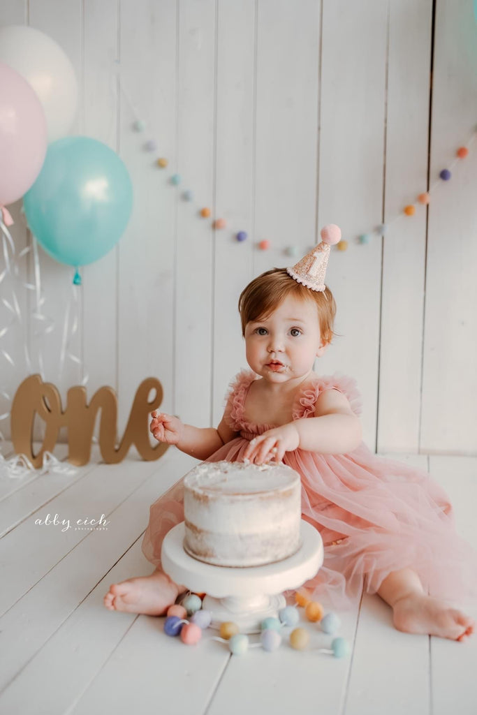 A baby in a pink dress and a Party Hat - Champagne Glitter with Blush Pom sits on the floor with a small cake in front of her. Colorful balloons and a “one” sign are in the background, along with a garland of hand crafted pom-poms, creating the perfect birthday accessory for her special day.