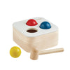 The Punch & Drop Bin is a classic cause-and-effect toy that includes a wooden box with three colored balls—red, blue, and yellow—and a sturdy mallet. Two of the balls are set in holes on top of the bin, while the yellow ball and mallet rest in front, inviting children to play.