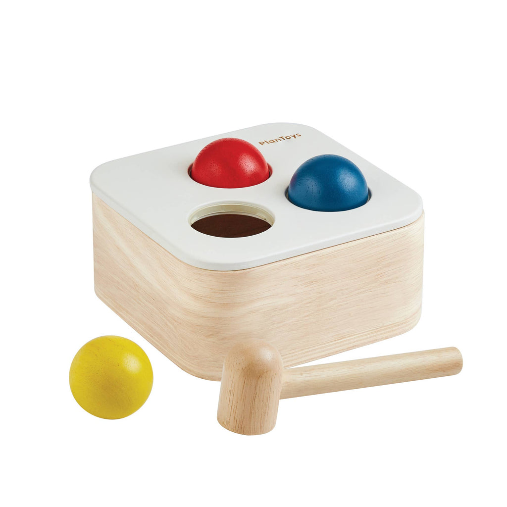 The Punch & Drop Bin is a classic cause-and-effect toy that includes a wooden box with three colored balls—red, blue, and yellow—and a sturdy mallet. Two of the balls are set in holes on top of the bin, while the yellow ball and mallet rest in front, inviting children to play.