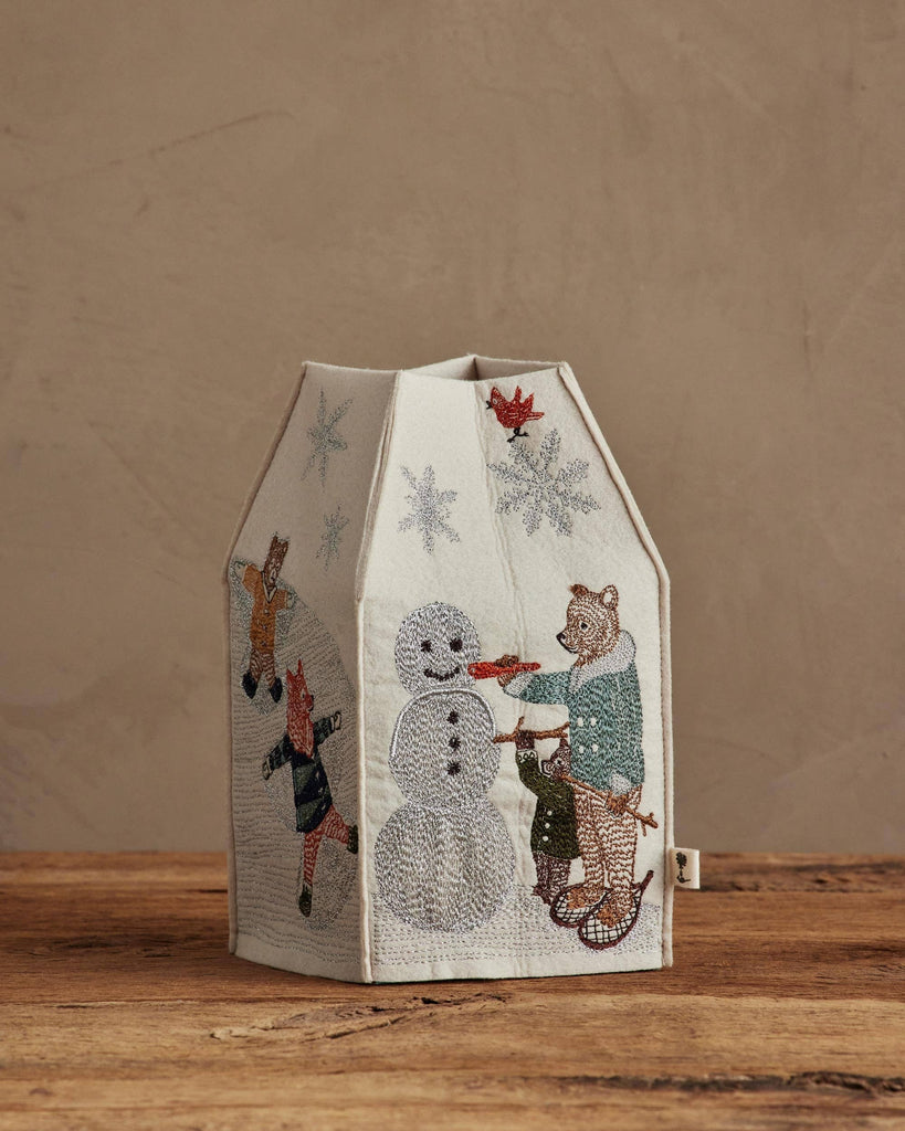 The Coral & Tusk Snow Day Tissue Box Cover features embroidered animals—a tiger, fox, and bear—building a snowman amid falling snowflakes, making it perfect whimsical winter decor for your wooden surface.