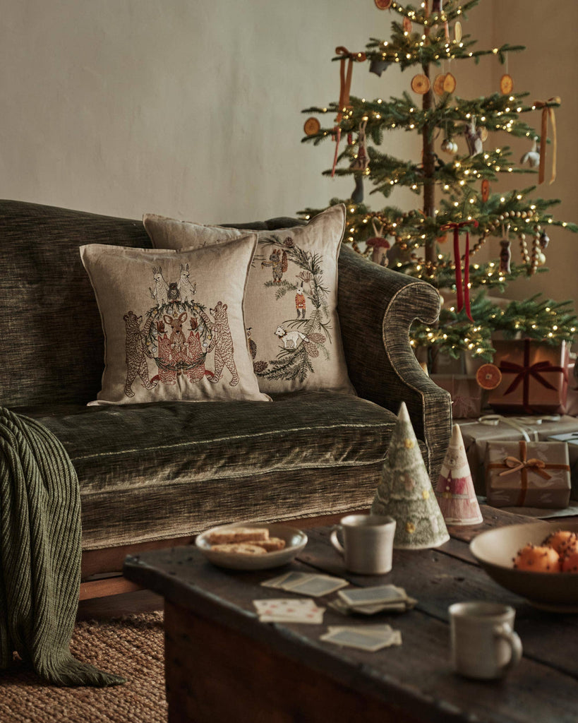 A cozy living room with a dark green sofa, a Coral & Tusk Tree Trimmers Wreath Pillow, an animal Christmas pillow, a green blanket, a wooden coffee table with snacks and drinks, and a decorated Christmas tree with lights and gifts.