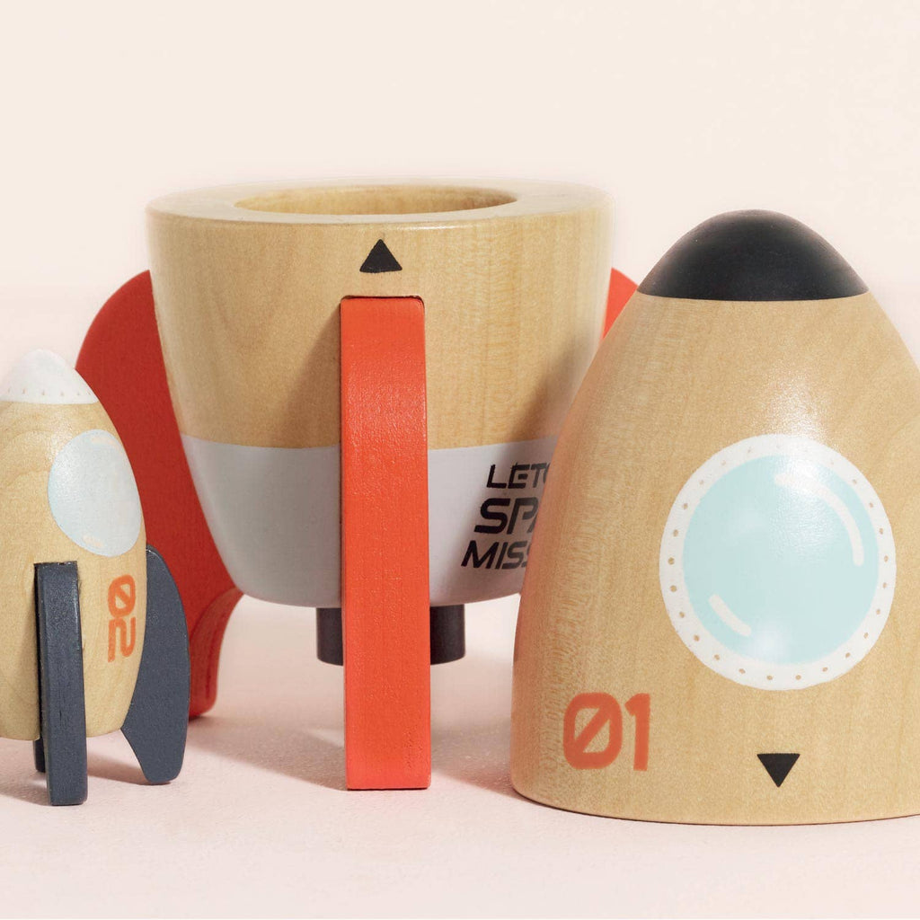 Two wooden toy rockets, inspired by the Magnetic Space Rocket Duo, feature detachable parts for creative play. Painted in beige, red, white, and black with window and number details, they're set against a light background.