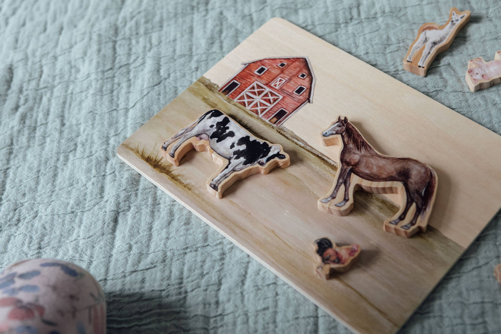 The Farm Animals Wooden Puzzle: A Tabitha Paige Puzzle features a cow and horse placed correctly on a barn-themed background, with other animal pieces nearby on a textured light green cloth—making it the perfect educational toy for toddlers.