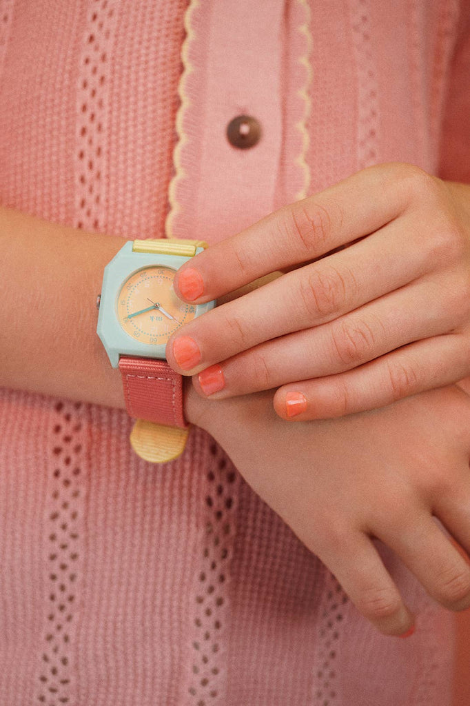 Wearing coral nail polish and a pink scalloped-edge knit top with a brown button, someone touches their Mini Kyomo Watch - Bubble Gum.