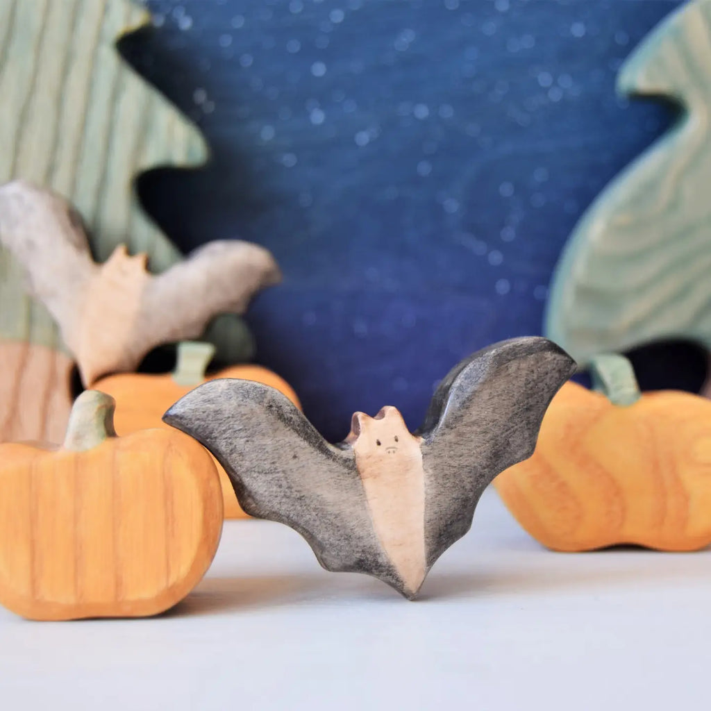 A scene with hand-crafted wooden carvings under a starry night sky backdrop. In the foreground, the Wooden Bat, a striking nocturnal animal, showcases its wings spread wide amidst pumpkins and trees made from sustainable hardwood. The pumpkins feature smooth, rounded shapes while the trees in the background exhibit green hues with natural textures.