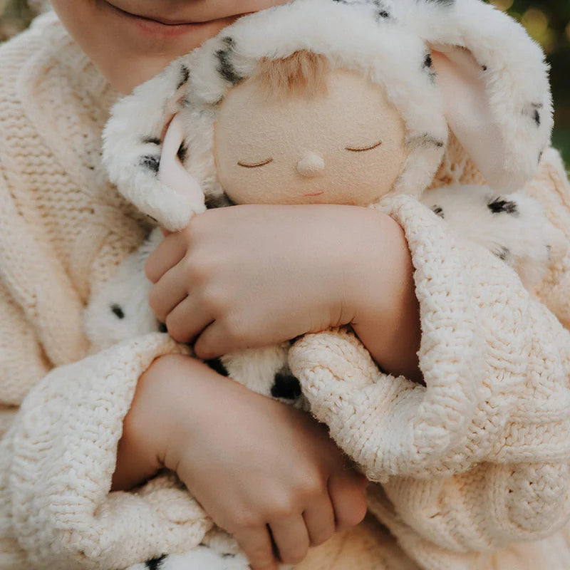 A child in a cream knit sweater hugs the Olli Ella Cozy Dinkum - Dog Domino, a plush toy dressed in a white spotted dog costume with long ears. The child's gentle smile is partially visible.
