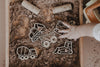 A child's hand reaches for the Construction Cutter Set - Eco Playdough Tool, featuring wooden construction vehicle toys like a dump truck and cement mixer, displayed on brown kinetic sand in a sensory play tray. This eco-friendly set encourages imaginative building and provides a hands-on sensory experience.