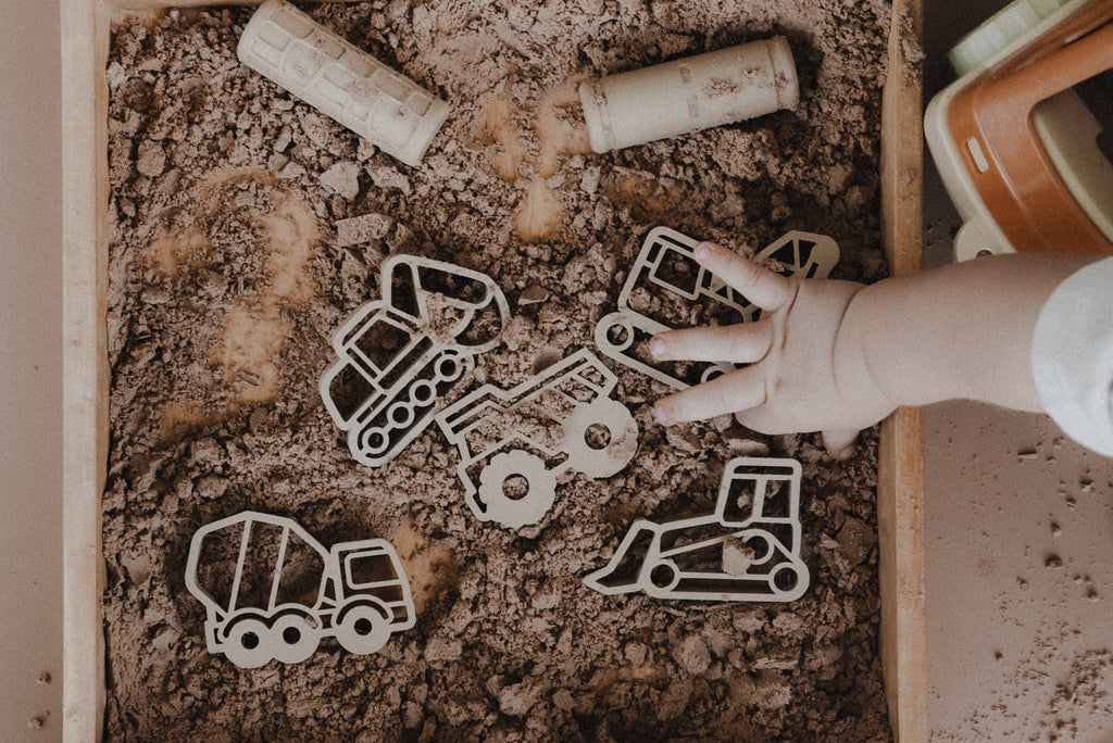 A child's hand reaches for the Construction Cutter Set - Eco Playdough Tool, featuring wooden construction vehicle toys like a dump truck and cement mixer, displayed on brown kinetic sand in a sensory play tray. This eco-friendly set encourages imaginative building and provides a hands-on sensory experience.