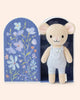 Cuddle + Kind Tiny Bailey the Mouse is a hand-knit, fair trade doll in light blue overalls, posed before a pastel box adorned with blue and purple flowers. This charmingly detailed toy makes a perfect gift.