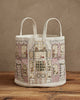 The Home for the Holidays Bucket is a white fabric tote bag with two handles, embroidered with a festive building decorated for Christmas, placed on a wooden surface against a beige wall.