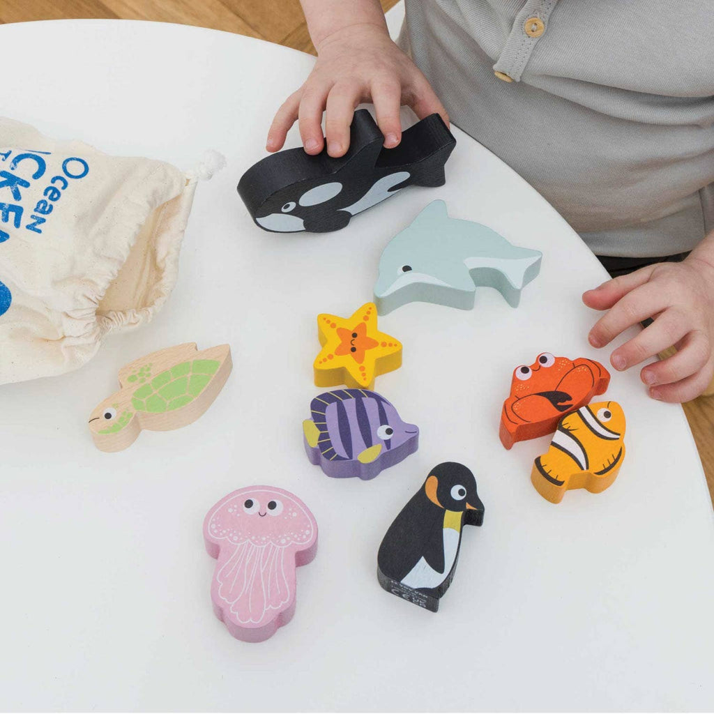 A child enjoys sensory play with the Ocean Life Stacking Animals & Bag, featuring colorful wooden sea animals like an orca, dolphin, and turtle on a white table. The labeled bag holds the ocean animal toy set nearby.