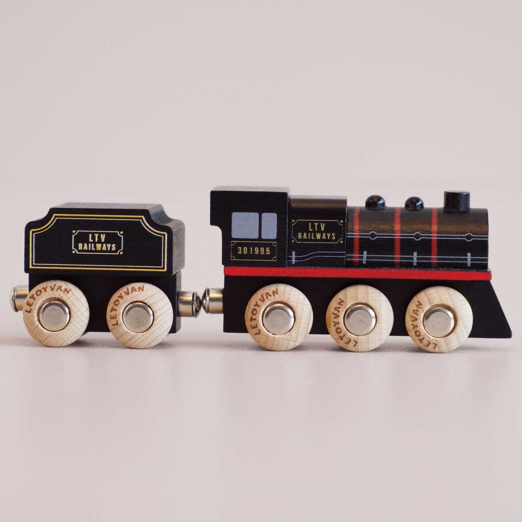 No. 49 GT295 Francis | Made in England: A charming wooden steam train engine and coal car, both painted black with gold and red detailing, stand connected on a light surface. Their light wood wheels are labeled “Le Toy Van,” capturing the nostalgic appeal of classic locomotives.