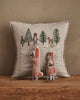 The Coral & Tusk Winter Foxes Pocket Pillow, featuring embroidered trees, a bear, and a deer, rests on wood with two plush fox toys—one upright, one doing a handstand—for charming animal nursery or winter fox decor.