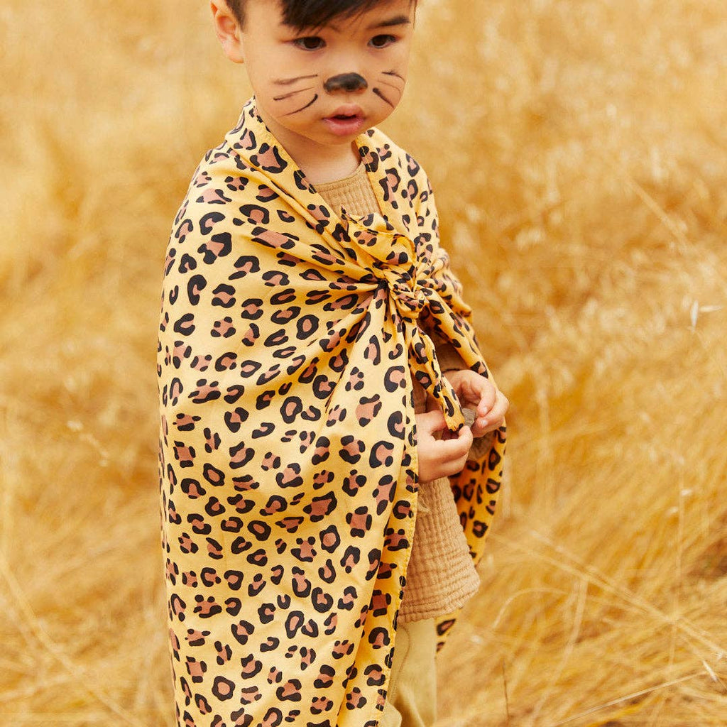 A young child stands in tall, dry grass, looking down while wearing Sarah's Silks Animal Print Playsilk - Cheetah as a cape and cat-like face paint, holding the pure silk cape closed at the front.
