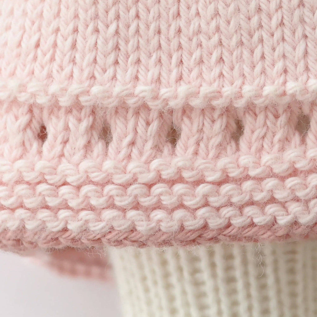 Close-up of Cuddle + Kind Clover the Bunny (Blossom) shows pale pink hand knit fabric with a decorative openwork row and ribbed cream section; handmade in Peru using fair trade practices.