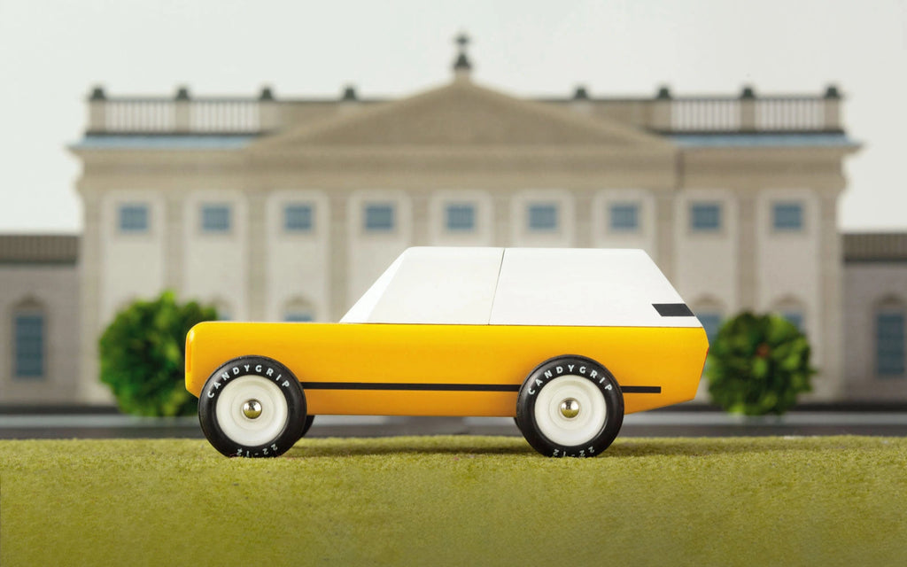 The Candylab Cotswold, a small yellow and white toy vehicle with black wheels, is displayed against a blurred backdrop of a neoclassical building. The car sits on a green surface, with parts of the building's ornate facade and symmetrical windows visible in the background.