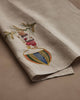 The Coral & Tusk Deck the Halls Bear Tea Towel is a beige linen towel embroidered with a bear in red shorts clinging to a tree branch on a striped, colorful ball—perfectly folded for festive holiday kitchen decor.