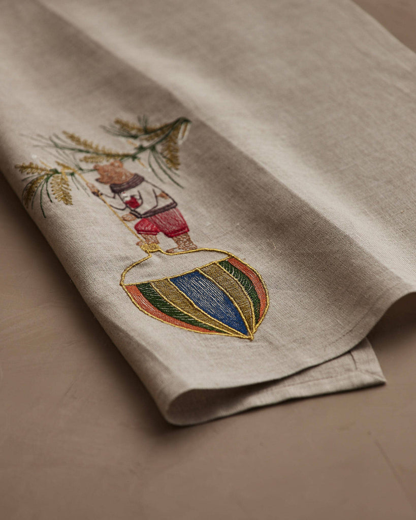 The Coral & Tusk Deck the Halls Bear Tea Towel is a beige linen towel embroidered with a bear in red shorts clinging to a tree branch on a striped, colorful ball—perfectly folded for festive holiday kitchen decor.